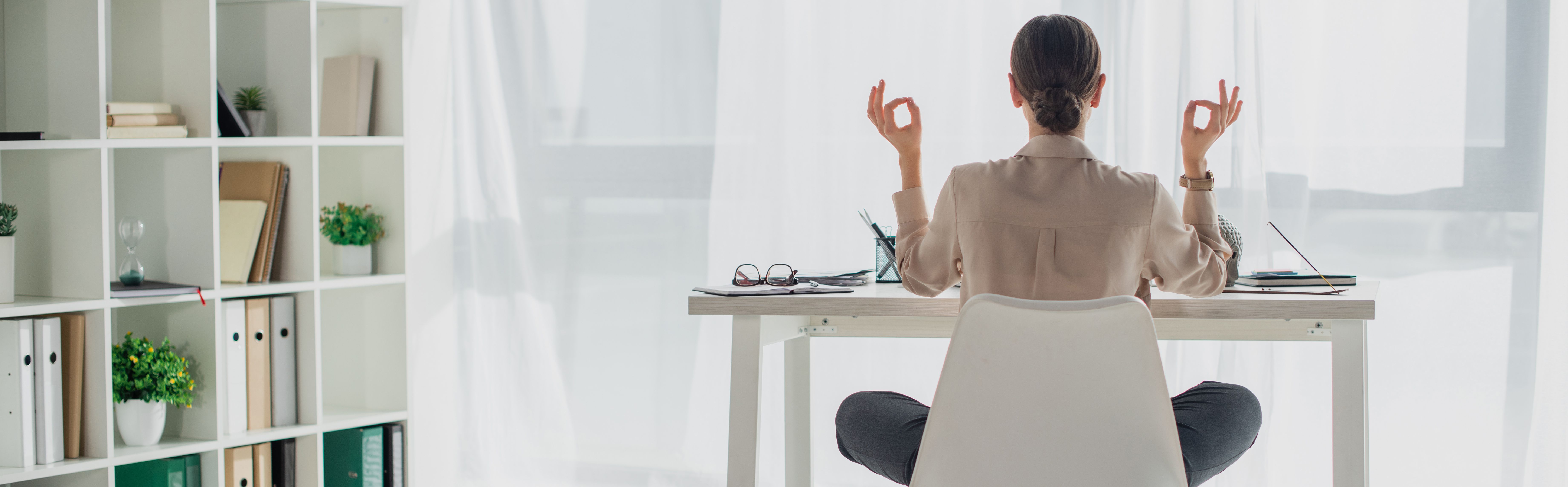 Person sitzt im Schneidersitz auf einem Stuhl, führt eine Meditationsgeste aus, während sie an einem Schreibtisch sitzt, umgeben von Büromaterialien und Pflanzen, beleuchtet von weichem, einfallendem Licht.