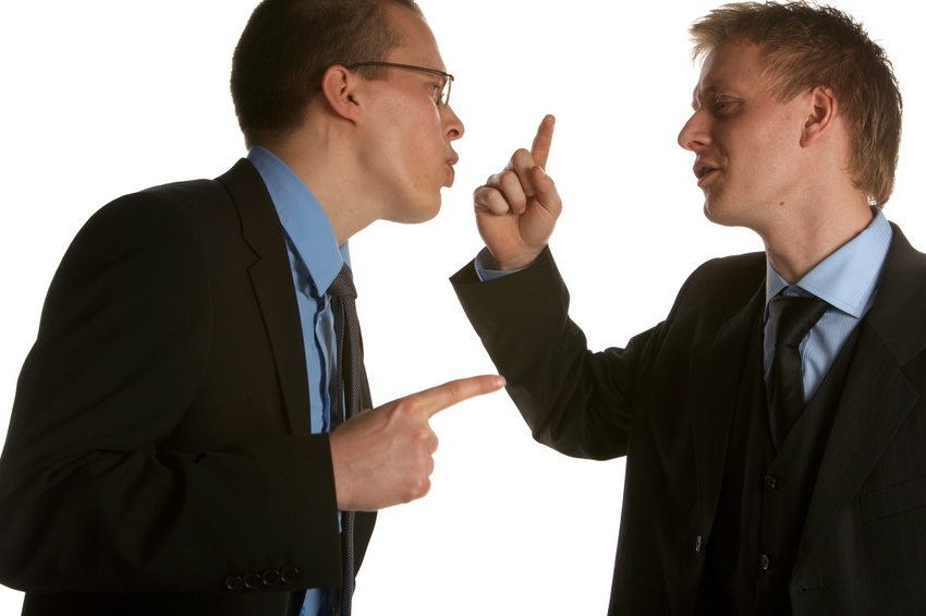 Zwei Männer in Anzügen stehen gegenüber, zeigen mit den Fingern aufeinander und diskutieren gestikulierend. Der Hintergrund ist schlicht weiß, was den Fokus auf die Interaktion lenkt.