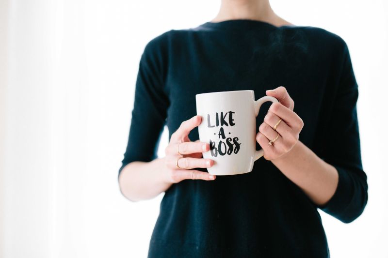 Eine Person in dunklem Oberteil hält eine weiße Tasse mit der Aufschrift „LIKE A BOSS“ in den Händen. Der Hintergrund ist unscharf und schlicht.