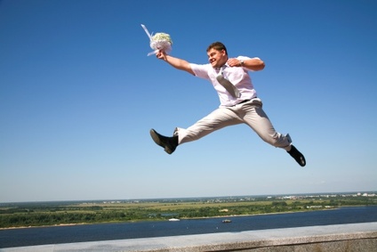 Ein Mann im Anzug springt dynamisch in die Luft, hält einen Blumenstrauß in der Hand und befindet sich vor einem klaren blauen Himmel mit einer weiten Landschaft im Hintergrund.