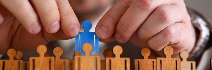 Eine Person platziert eine blaue Figur zwischen mehrere braune Holzfiguren, die aufrecht stehen. Hintergrund ist verschwommen, die Hände sind im Fokus und vermitteln eine Auswahl- oder Entscheidungsaktion.