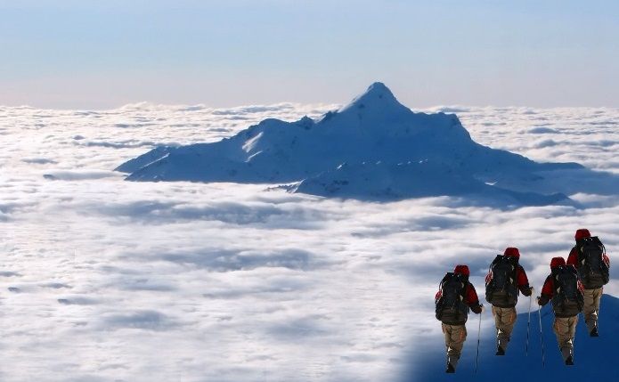 Bergsteiger mit roten Rucksäcken wandern eine schneebedeckte Fläche entlang, während im Hintergrund ein schneebedeckter Berg majestätisch aus einer Wolkendecke herausragt, die den Himmel bedeckt.