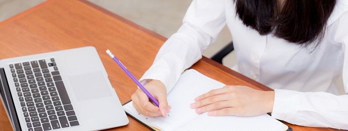 Person in weißem Hemd schreibt mit einem lila Stift in ein Notizbuch. Auf dem Holztisch steht ein offenes Laptop. Die Umgebung wirkt wie ein Büroumfeld.