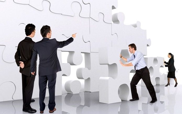 Personen in Geschäftskleidung interagieren mit übergroßen Puzzleteilen; einige schieben die Teile zusammen, während andere das Puzzle inspizieren. Der Hintergrund ist neutral, was den Fokus auf die Teamarbeit legt.