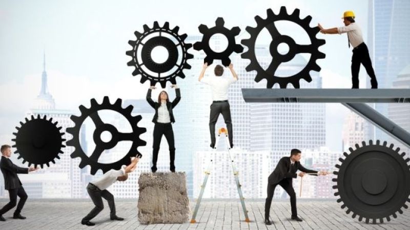 Zahnräder in verschiedenen Größen werden von mehreren Personen in Geschäftskleidung und einem Bauarbeiter zusammengefügt. Sie stehen auf Leitern und einem Podest vor einer Stadtsilhouette, um ein komplexes Mechanismus zu schaffen.