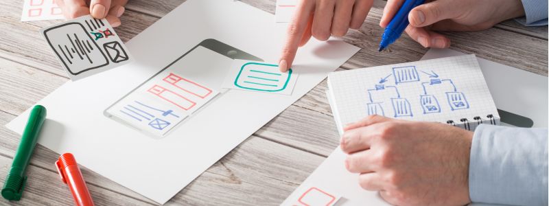 Mehrere Hände arbeiten an gedruckten Design- und Konzeptzeichnungen auf einem Holztisch, mit Markern und Stiften, während technische Skizzen und Diagramme markiert werden.