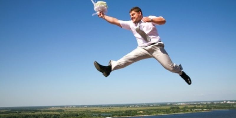 Ein Mann im Anzug springt dynamisch in die Luft, hält einen Blumenstrauß in der Hand und befindet sich vor einem klaren blauen Himmel mit einer weiten Landschaft im Hintergrund.
