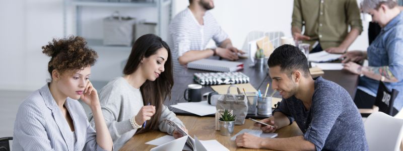 Personen arbeiten gemeinsam an einem langen Tisch, konzentriert auf Laptops und Tablets. Im Hintergrund stehen Ablagen und Regale, die eine moderne Büroatmosphäre schaffen. Keine sichtbaren Textelemente.