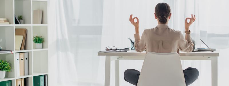 Person sitzt im Schneidersitz auf einem Stuhl, führt eine Meditationsgeste aus, während sie an einem Schreibtisch sitzt, umgeben von Büromaterialien und Pflanzen, beleuchtet von weichem, einfallendem Licht.