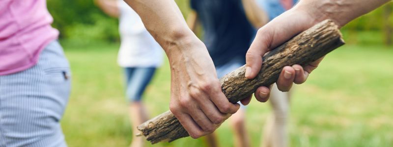 Zwei Hände greifen gemeinsam einen hölzernen Stab, wobei der Hintergrund verschwommen ist und andere Personen leicht zu erkennen sind. Der Kontext deutet auf eine Aktivität im Freien hin.
