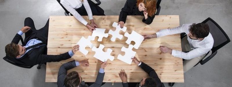 Gruppe von Menschen sitzt an einem großen Holztisch und setzt mehrere Puzzleteile zusammen.