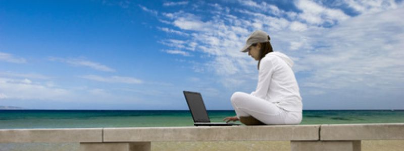 Eine Person sitzt in weißer Kleidung und Mütze auf einer Betonbank am Strand und arbeitet am Laptop, während der blaue Himmel mit Wolken die Szenerie umrahmt.