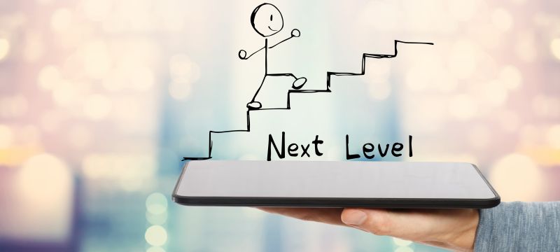 Ein gezeichneter Strichmännchen steigt eine Treppe empor, mit der Beschriftung „Next Level“ darunter. Die Treppe wird auf einem in einer Hand gehaltenen Tablet dargestellt. Hintergrund unscharf.